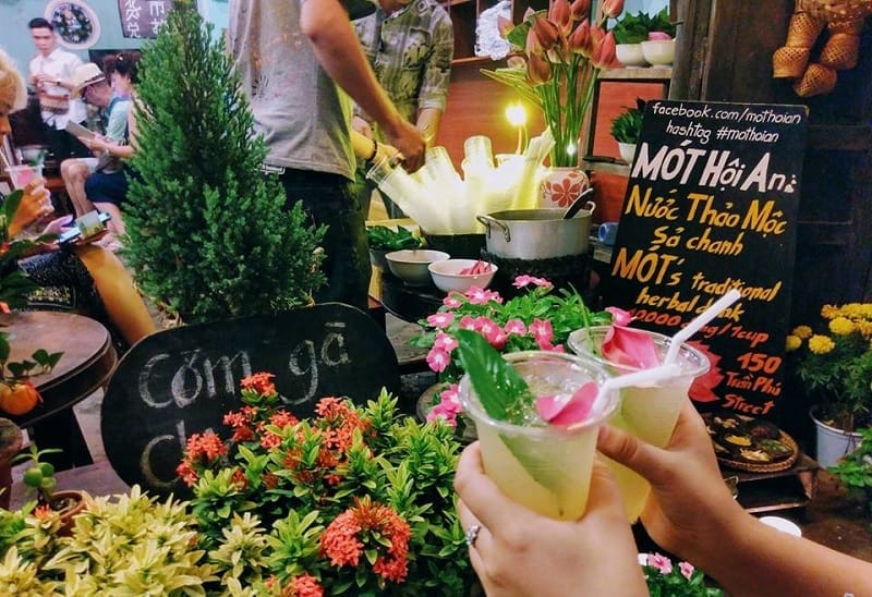 Nếu bạn đang muốn tìm một địa chỉ ăn vặt Hội An đậm chất phố cổ, Mót Hội An chắc chắn là điểm dừng chân lý tưởng. Nơi đây nổi tiếng với món nước thảo mộc thanh mát, mang hương sen dịu nhẹ, quyện cùng vị chanh và sả thoang thoảng. Chỉ với 10.000VNĐ, bạn đã có thể cầm trên tay một ly nước giải khát mát lạnh, được chăm chút bằng chiếc lá tươi và cánh hoa sen nhỏ xinh – vừa ngon miệng lại vừa đẹp mắt, rất “Hội An”.