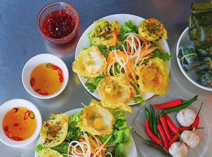 Quán bánh căn Cô Bé – một điểm đến ăn vặt quen thuộc tại Hội An – ghi điểm không chỉ bởi không gian sạch sẽ, gọn gàng mà còn bởi những đĩa bánh căn chất lượng, hấp dẫn từ hương đến vị. Mỗi chiếc bánh nóng hổi có lớp vỏ giòn rụm, trứng béo thơm, ăn kèm với đồ chua, chả và nem đậm đà, tạo nên hương vị hài hòa khó quên ngay từ miếng đầu tiên.
